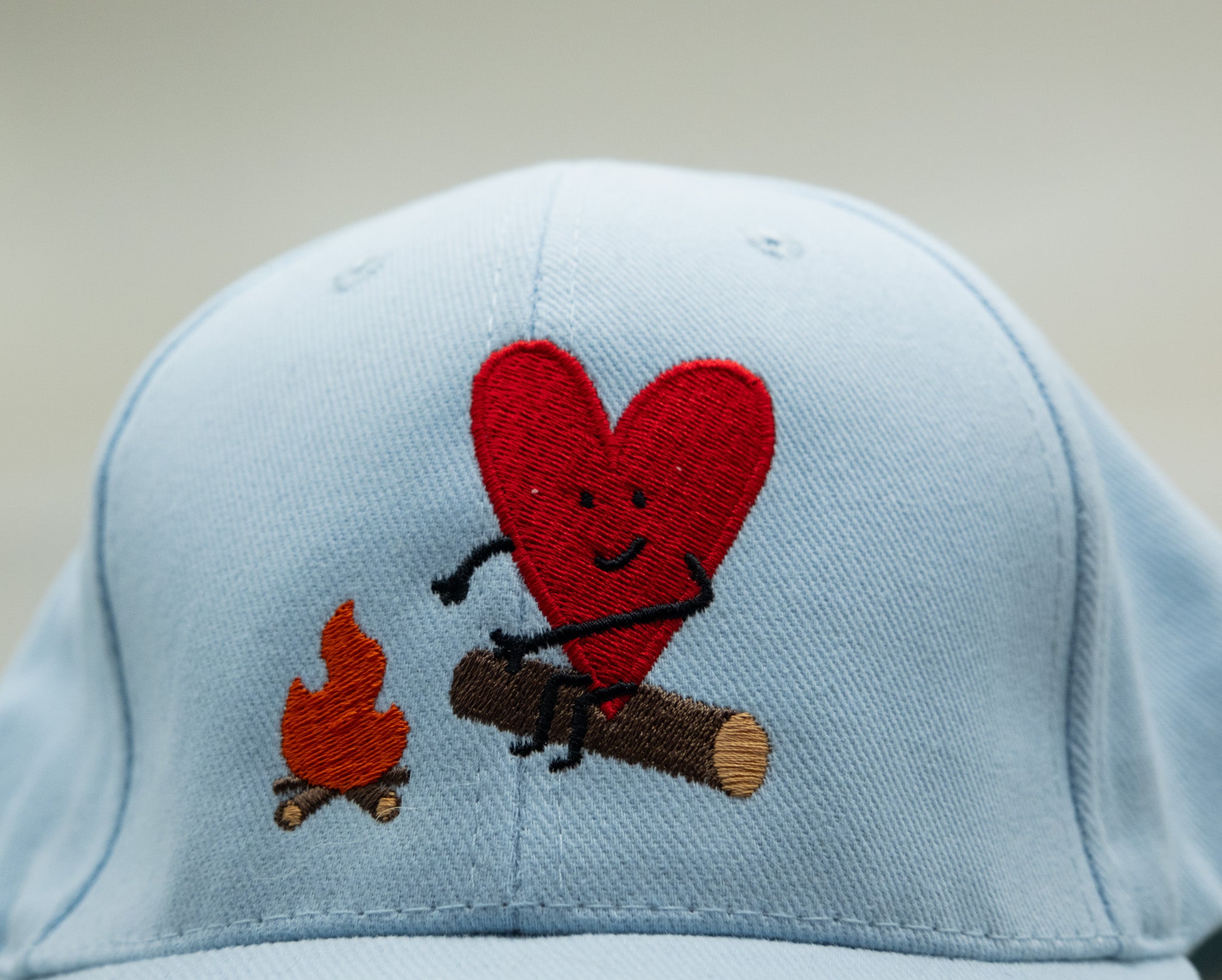 Light blue cap with a red heart and black stick figure near a campfire on a beige background