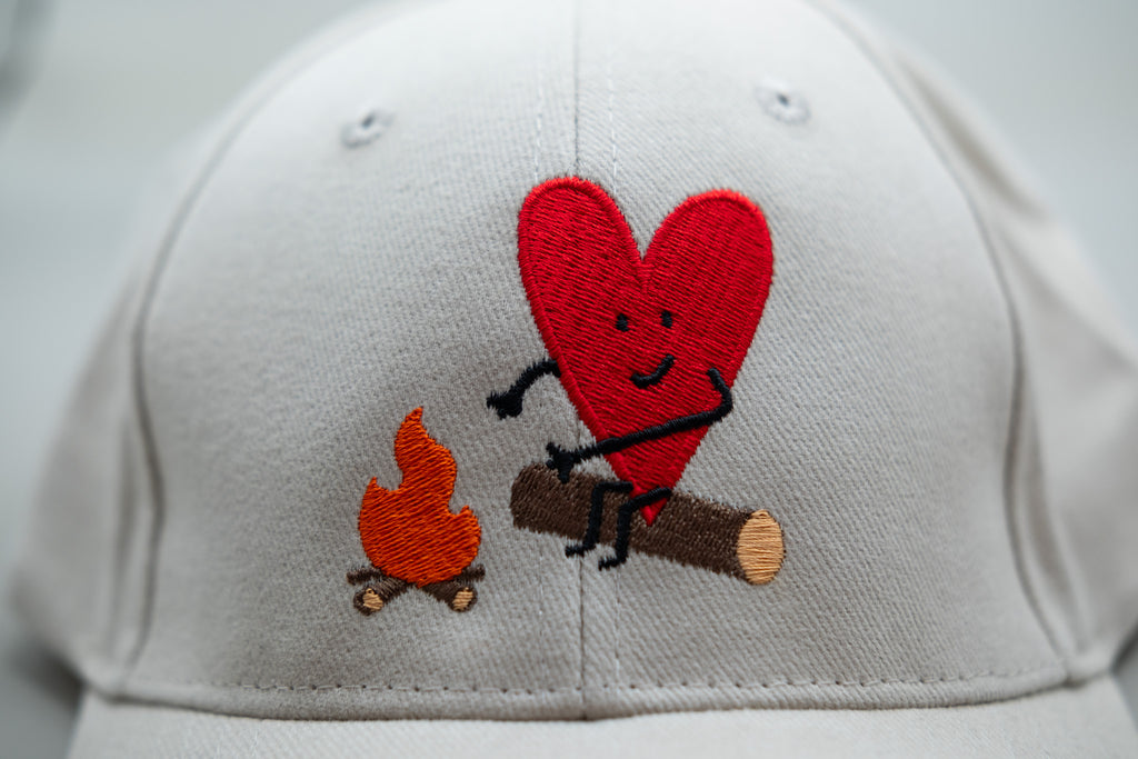 White cap with a red heart and campfire design on a white background