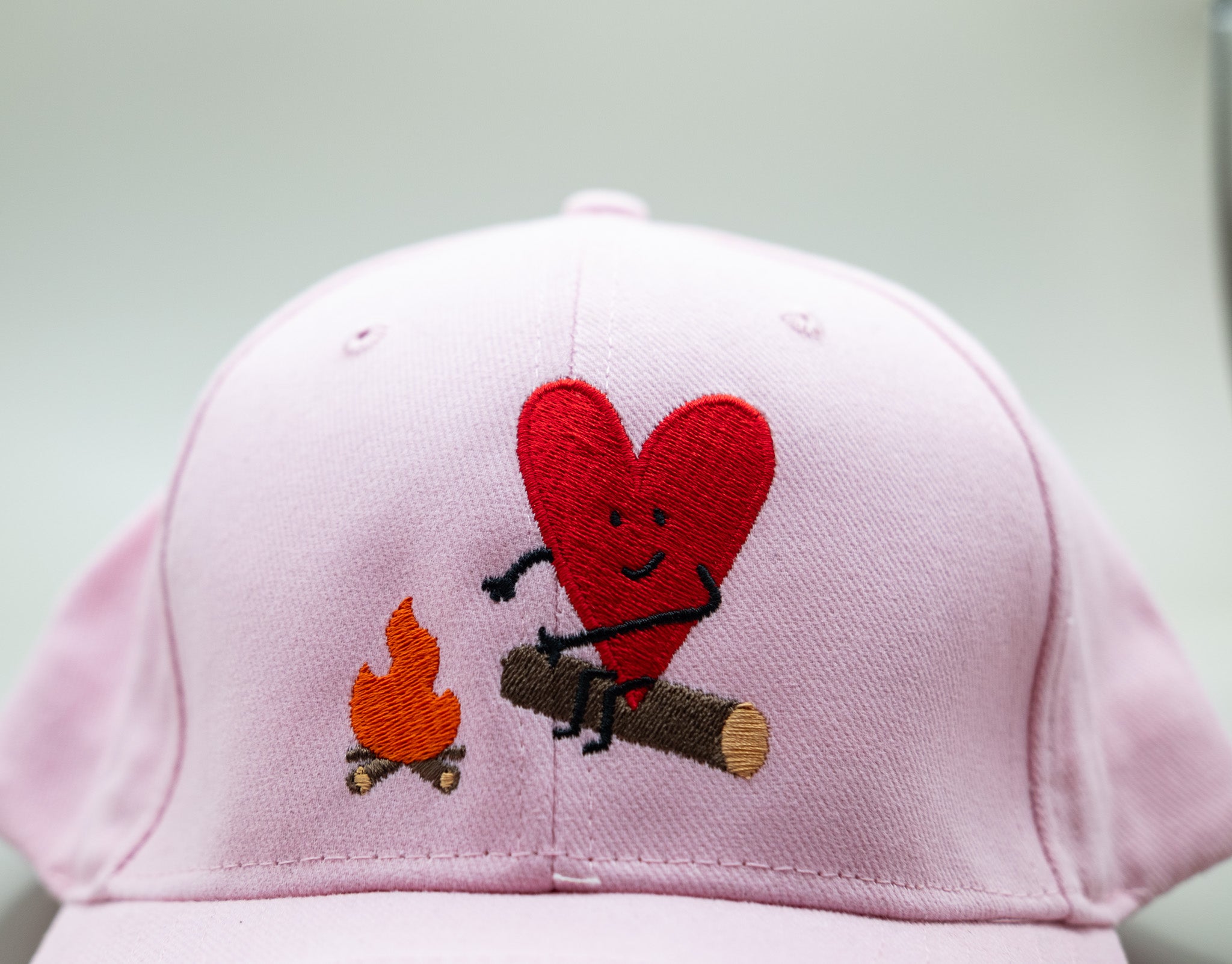 Pink cap with a red heart, black stick figure, orange campfire, and brown log on a white background