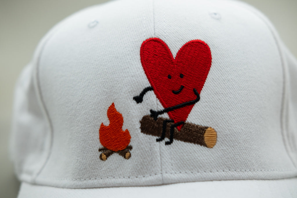 White cap with a red heart and campfire design on a plain background