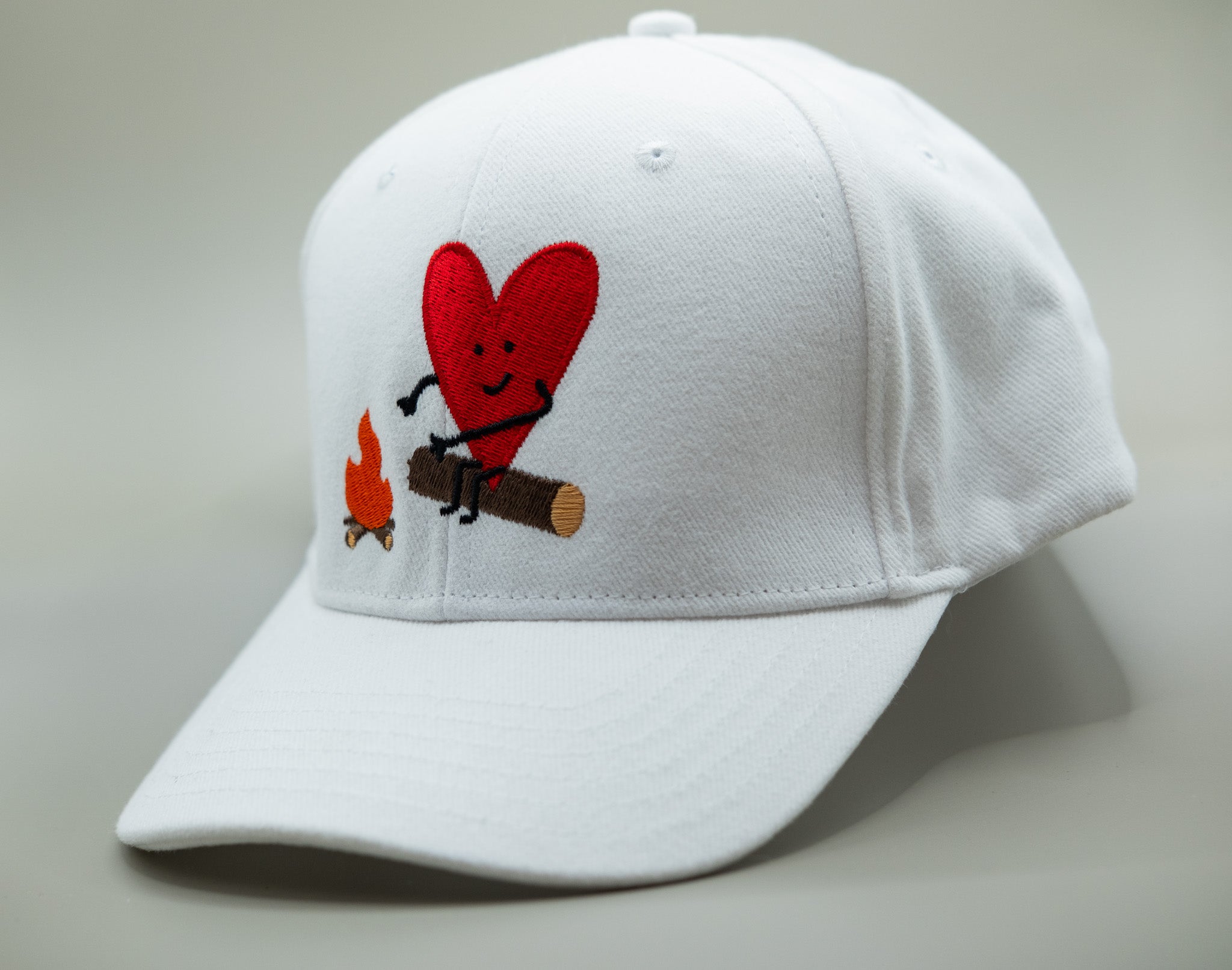 White cap with a red heart and camping-themed design on a light gray background